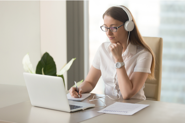 Focused attentive woman in headphones sits at desk with laptop, looks at screen, makes notes, learns foreign language in internet, online study course, self-education on web, consults client by video